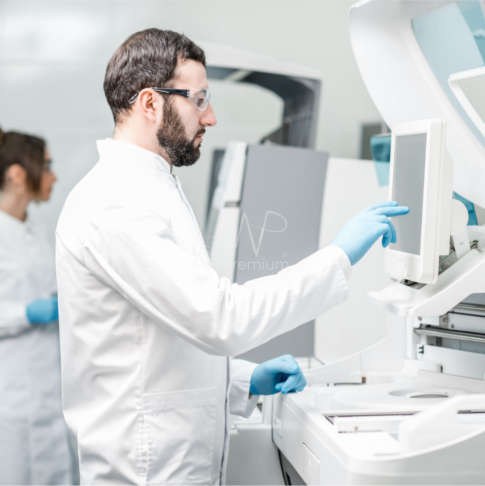 Lab technician inspecting a blood collection tube beside racks of specimen tubes while a colleague loads a diagnostic analyzer